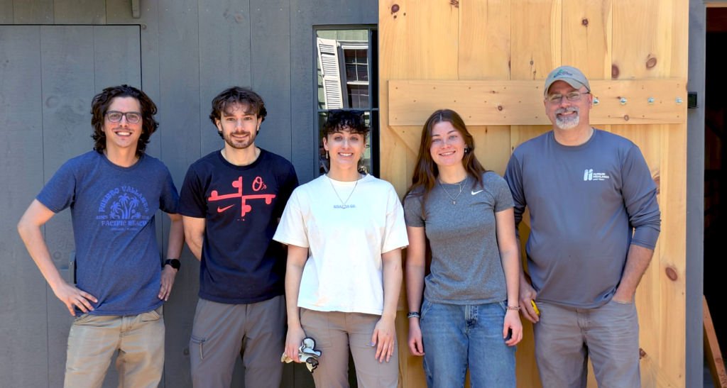 A group of people stand in front of a gray barn. The group contains the staff and interns of the Mianus River Gorge