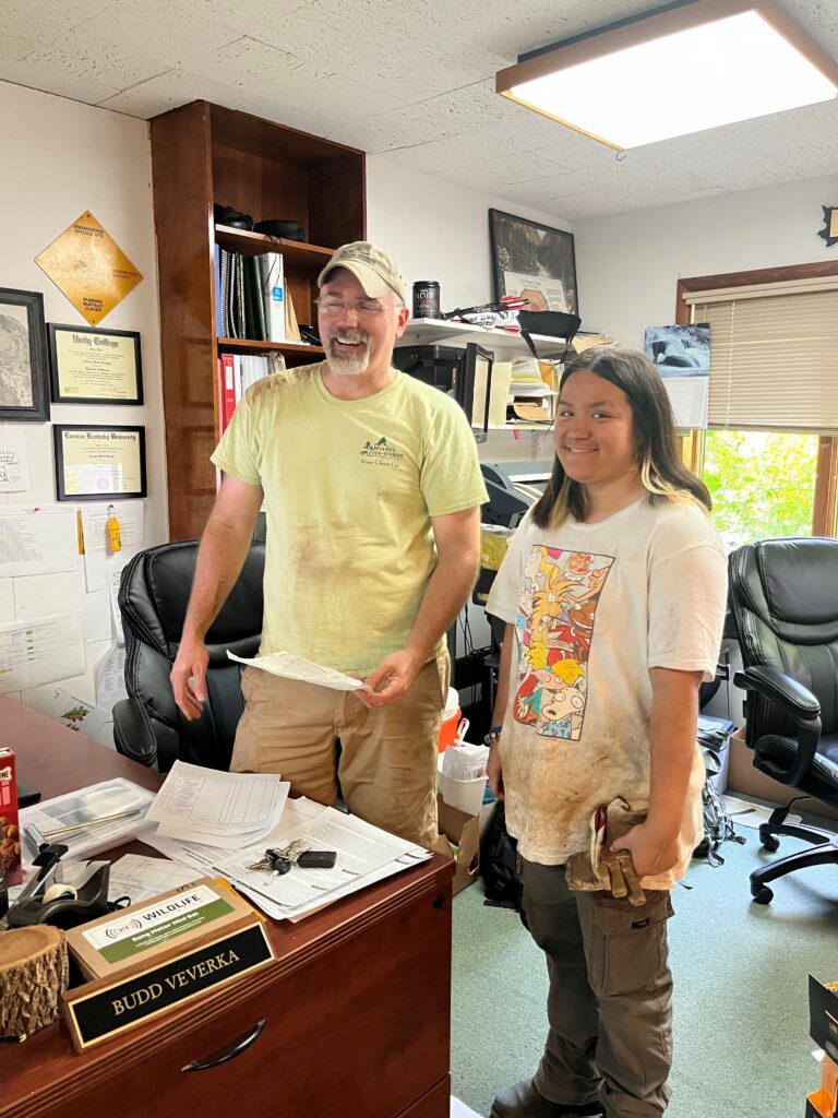 Staff and student, a man in a yellow t-shirt with a woman in a graphic t-shirt, stand in a crowded office